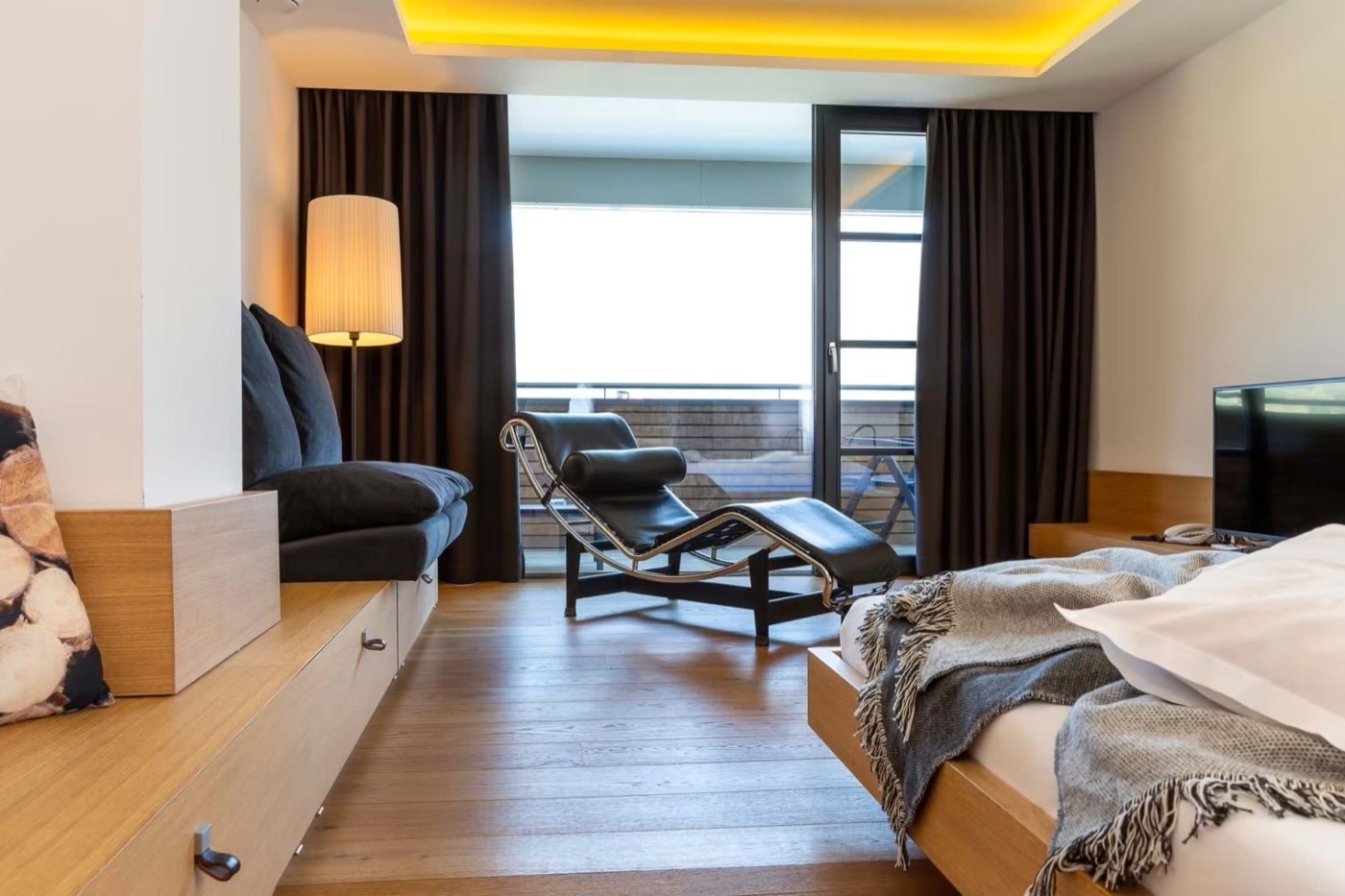 Doppelzimmer im Pazeider mit Eichenboden, schwarzer Liege aus Leder und Stahl vor der bodentiefen Fensterfront, rechts das Bett mit grauer Fransendecke, dahinter die Schiebetür zum Balkon