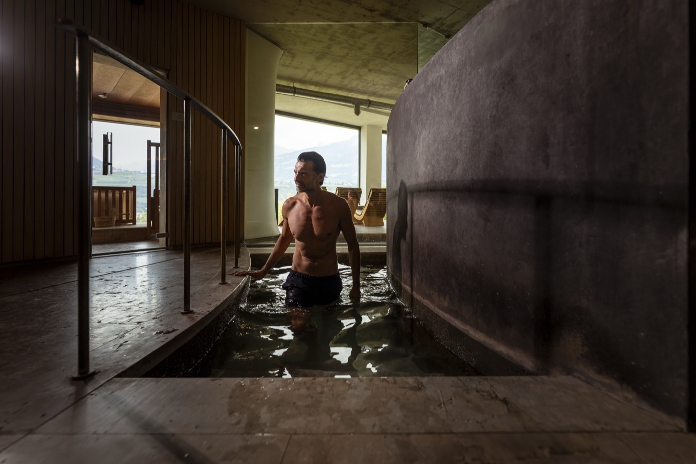 Ein Mann steht in einem in Stein gefassten Tauchbecken im Spa und hält sich am Handlauf, im Hintergrund ein Fenster mit Blick ins Tal